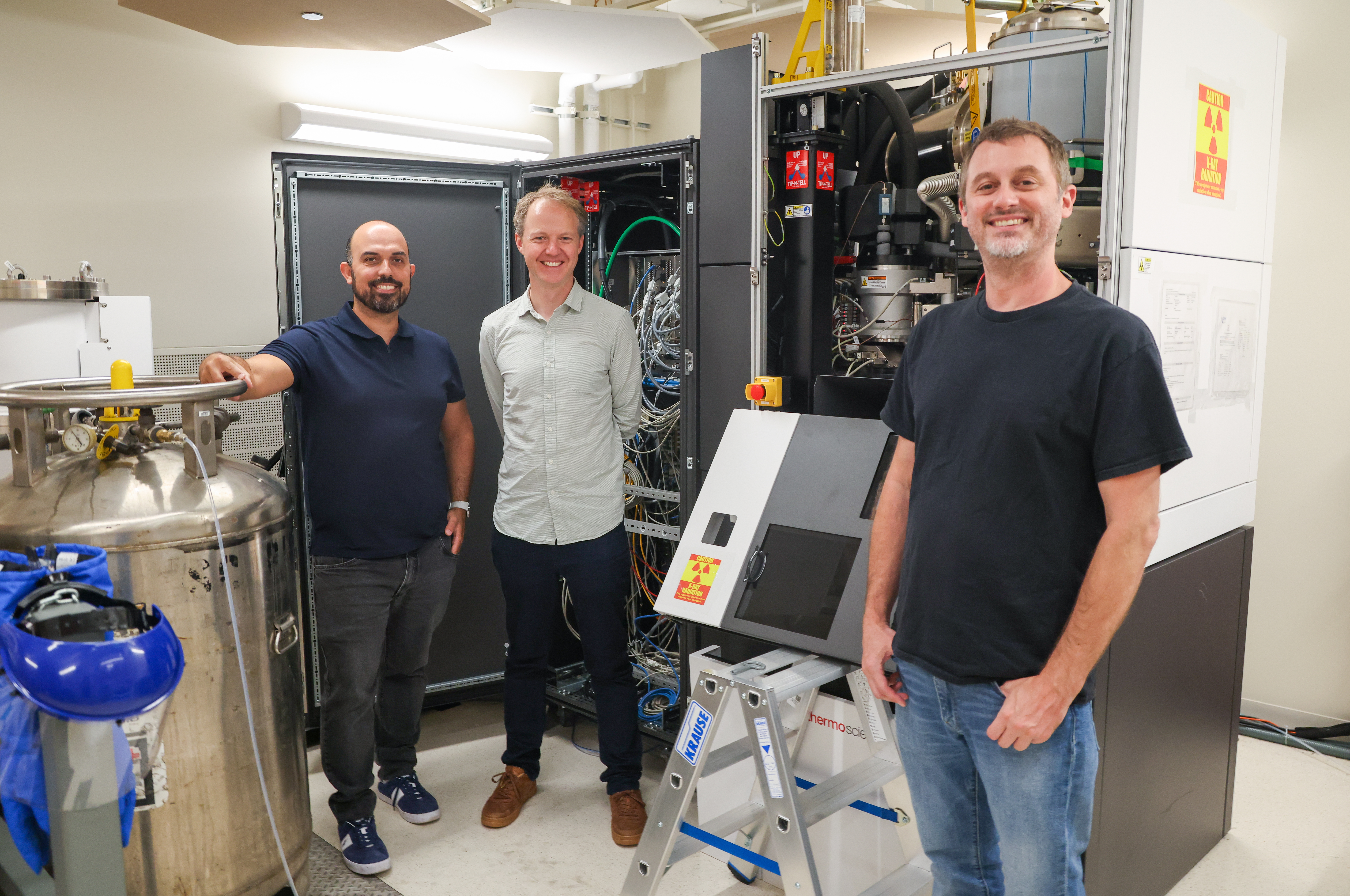 University staff and visitor standing in front of large electronic research apparatus